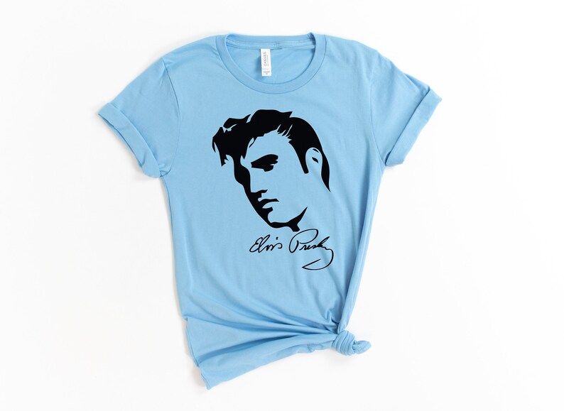 May include: Light blue t-shirt with a black silhouette of Elvis Presley's face and his signature "Elvis Presley".