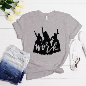 Peut inclure: Un t-shirt gris chin&eacute; avec une silhouette noire de trois femmes en robes et le texte "work" en &eacute;criture noire.