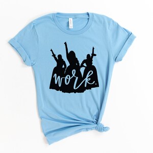 Peut inclure: T-shirt bleu clair avec une silhouette noire de trois femmes en robes et le mot "work" &eacute;crit en cursive noire.