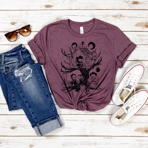 May include: A maroon t-shirt with a black graphic print of a treehouse with several figures standing around it. The figures are all wearing different outfits and have different hairstyles. There are also some balloons and a crescent moon in the graphic. The t-shirt is laid out on a white wooden surface with a pair of jeans, sunglasses, and a pair of white sneakers.