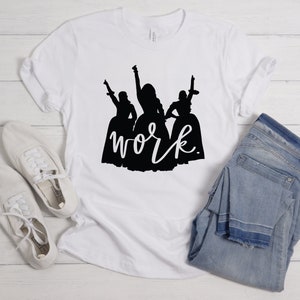 Peut inclure: T-shirt blanc avec une silhouette noire de trois femmes en robes et le mot "work" &eacute;crit en cursive en dessous.