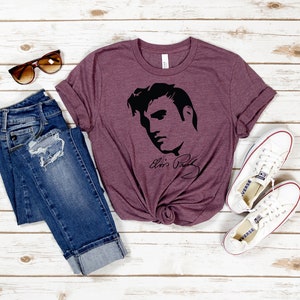 May include: A maroon t-shirt with a black silhouette of Elvis Presley's face and signature. The shirt is paired with blue jeans and white sneakers.