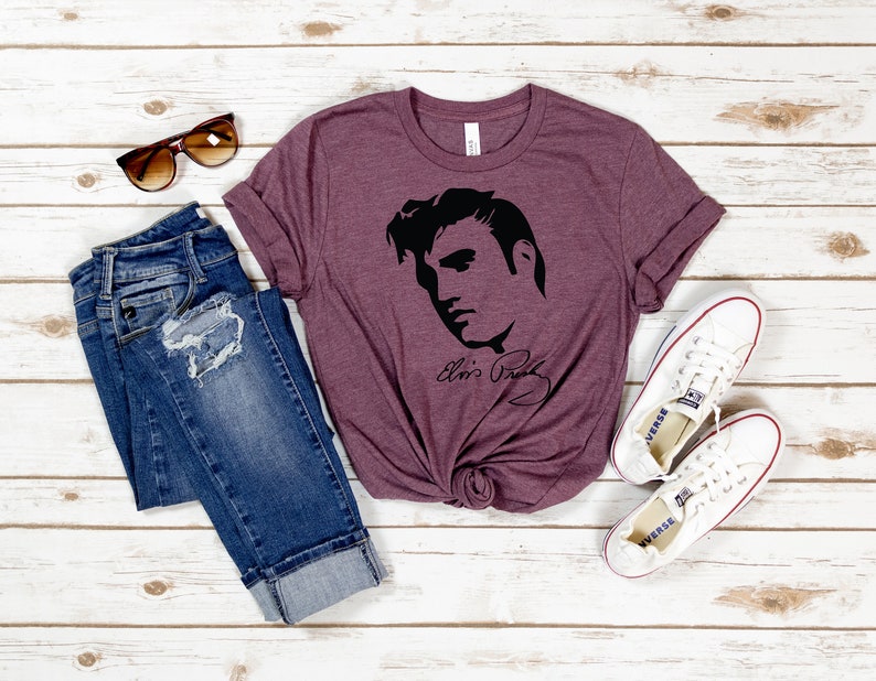 May include: A maroon t-shirt with a black silhouette of Elvis Presley's face and signature. The shirt is paired with blue jeans and white sneakers.