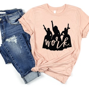 Peut inclure: Un t-shirt couleur p&ecirc;che avec une silhouette noire de trois femmes en robes et le mot "work" &eacute;crit en dessous.