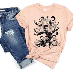 May include: A peach-colored t-shirt with a black graphic of horror movie characters in a treehouse. The characters include Freddy Krueger, Jason Voorhees, Michael Myers, and others.
