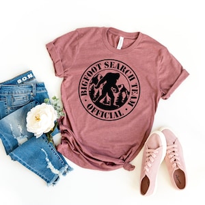 May include: A mauve t-shirt with a black circle graphic that says "Bigfoot Search Team Official" with a silhouette of Bigfoot walking in the woods.