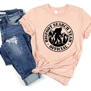 May include: A peach-colored t-shirt with a black circle graphic that says "Bigfoot Search Team Official" with a silhouette of Bigfoot walking in the woods.