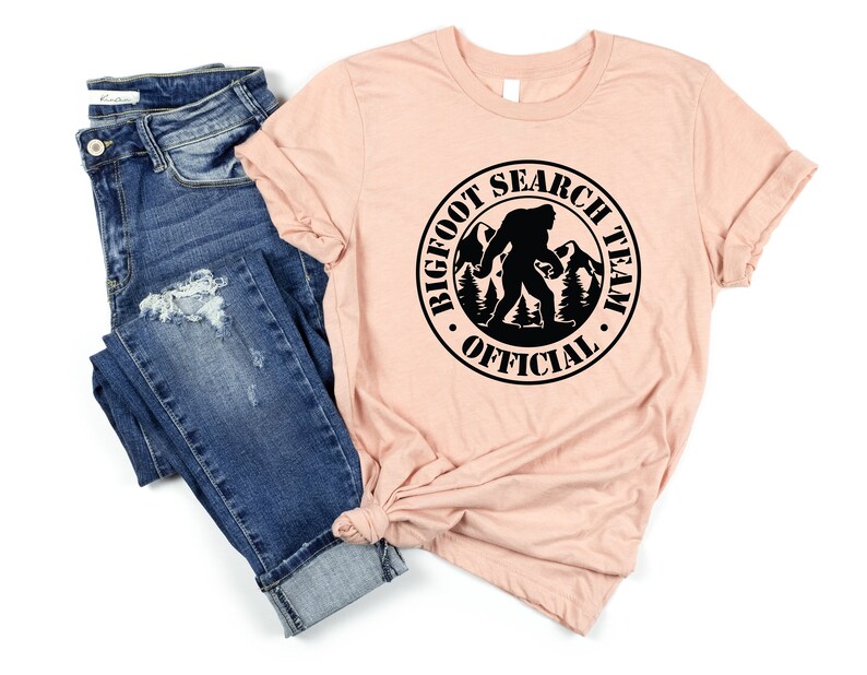 May include: A peach-colored t-shirt with a black circle graphic that says "Bigfoot Search Team Official" with a silhouette of Bigfoot walking in the woods.
