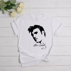May include: White t-shirt with a black silhouette of Elvis Presley's face and his signature "Elvis Presley".