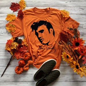 May include: Orange t-shirt with a black silhouette of Elvis Presley's face and signature. The shirt is surrounded by fall-themed decorations, including pumpkins and fall leaves.