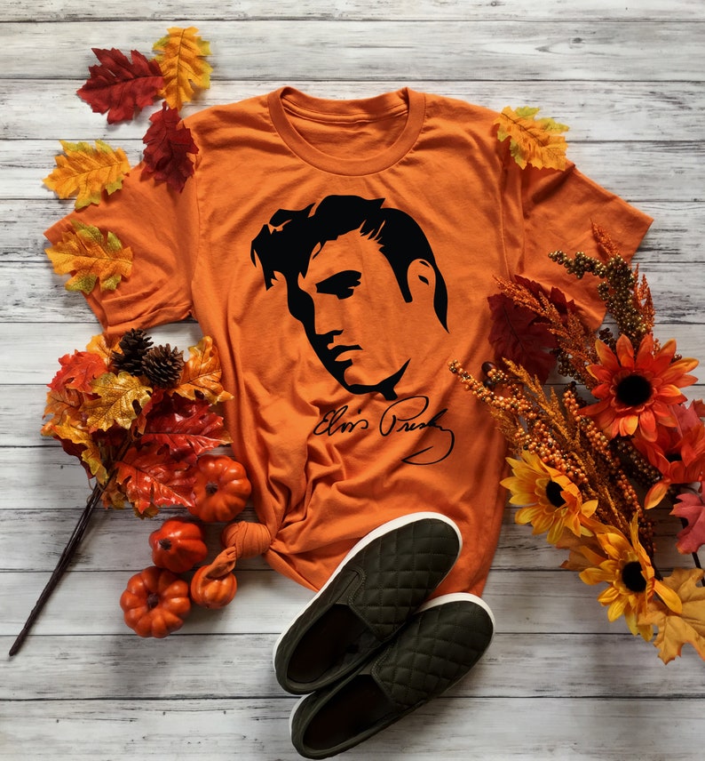 May include: Orange t-shirt with a black silhouette of Elvis Presley's face and signature. The shirt is surrounded by fall-themed decorations, including pumpkins and fall leaves.
