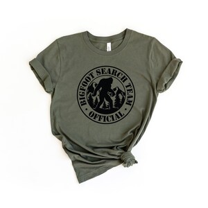 May include: A green t-shirt with a black circle graphic that says "Bigfoot Search Team Official" with a silhouette of Bigfoot walking in the mountains.