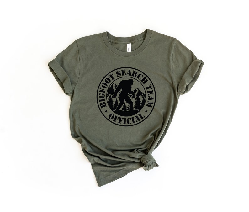May include: A green t-shirt with a black circle graphic that says "Bigfoot Search Team Official" with a silhouette of Bigfoot walking in the mountains.