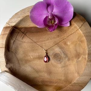 Könnte beinhalten: Eine goldfarbene Halskette mit einem tropfenförmigen, roten Edelsteinanhänger. Die Kette liegt auf einer Holzschale, neben einem weißen Kristall und einer leuchtend lila Orchidee. Die Anordnung ist schlicht und elegant.