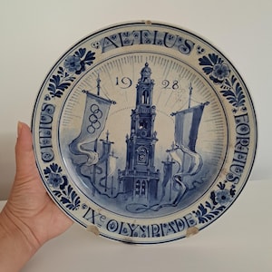 Może przedstawiać: Niebiesko-biały ceramiczny talerz z centralną ilustracją wieży, flagami i pierścieniami olimpijskimi. Talerz jest ozdobiony kwiatowymi wzorami oraz napisem "IXe OLYMPIADE" i "1928".