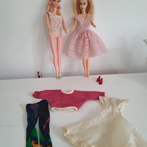 May include: Two vintage dolls with blonde hair and various outfits. One doll wears a pink dress, and the other wears a polka dot top. Accessories include red shoes, a pink sweater, patterned leggings, and a white dress.
