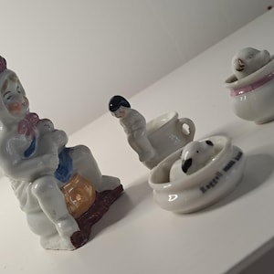 May include: A collection of small, white porcelain figurines. One figurine depicts a seated figure holding a baby, another is a figure in a teacup, and two others are in small bowls. The figurines have painted details in pink, blue, and brown.