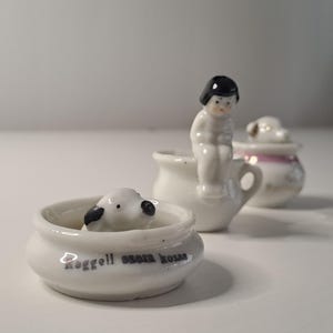 Pode incluir: Uma coleção de pequenas figuras de porcelana branca. Uma figura mostra uma criança sentada numa panela pequena. Outra mostra uma ovelha numa tigela com texto. Uma terceira figura é uma panela pequena com tampa. As figuras estão numa superfície branca.