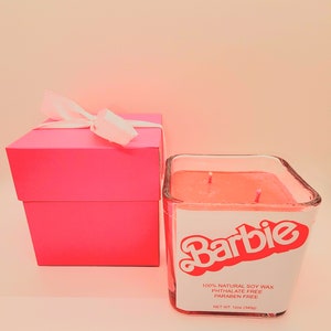 May include: A pink gift box with a white ribbon sits next to a square glass candle. The candle has a pink wax and a label with the word "Barbie" in red. The label also states the candle is 100% natural soy wax and 12oz (340g).