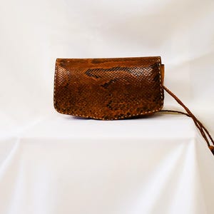 May include: A small, brown, rectangular clutch bag with a textured snakeskin pattern. The bag has a matching brown wrist strap. The background is a white fabric.