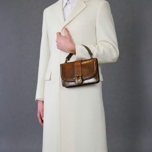 May include: A long, cream-coloured overcoat with a classic design. A brown leather handbag with a top handle and buckle closure is held. The handbag features a gold-coloured metal clasp.
