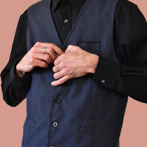 Puede incluir: Un chaleco a rayas azul y blanco con una camisa abotonada negra debajo. El chaleco tiene un bolsillo en el lado izquierdo y tres botones en la parte delantera.