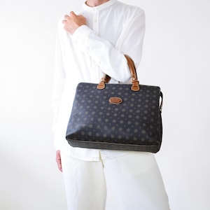 Könnte beinhalten: Eine dunkelbraune, rechteckige Handtasche mit einem sich wiederholenden, goldfarbenen, kreisförmigen Monogrammmuster auf der Oberfläche. Die Tasche verfügt über zwei hellbraune Ledergriffe mit goldfarbenen Beschlägen und ein ovales Markenlogo.