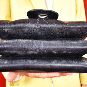 May include: A black leather handbag with a unique, layered design. The bag has a rounded top flap with a silver clasp. The bag is held against a red background, with a yellow blazer visible in the background.