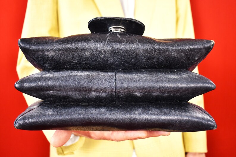 May include: A black leather handbag with a unique, layered design. The bag has a rounded top flap with a silver clasp. The bag is held against a red background, with a yellow blazer visible in the background.
