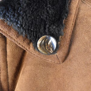 May include: Close-up of a brown suede jacket with a black faux fur collar and a silver button.