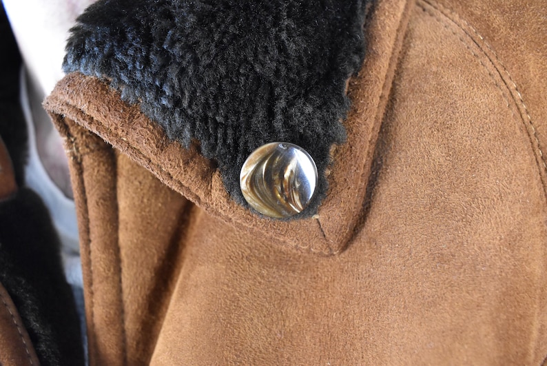 May include: Close-up of a brown suede jacket with a black faux fur collar and a silver button.