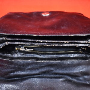 May include: Black leather clutch with a fold-over design. The clutch has a zipper closure and a silver-toned snap. The interior is visible, revealing a black lining. The bag is open, displaying its layered construction.