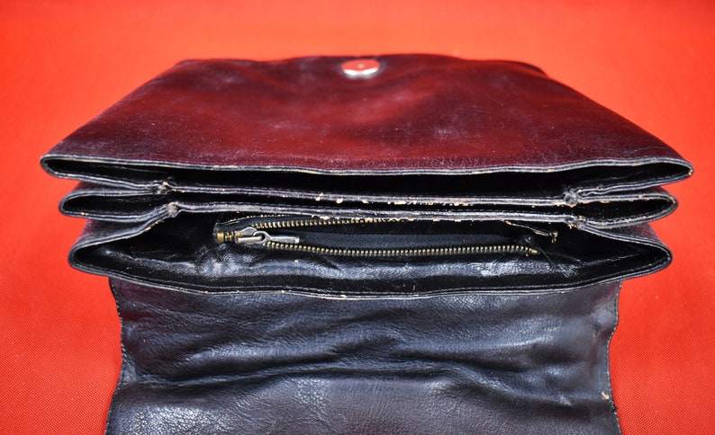 May include: Black leather clutch with a fold-over design. The clutch has a zipper closure and a silver-toned snap. The interior is visible, revealing a black lining. The bag is open, displaying its layered construction.