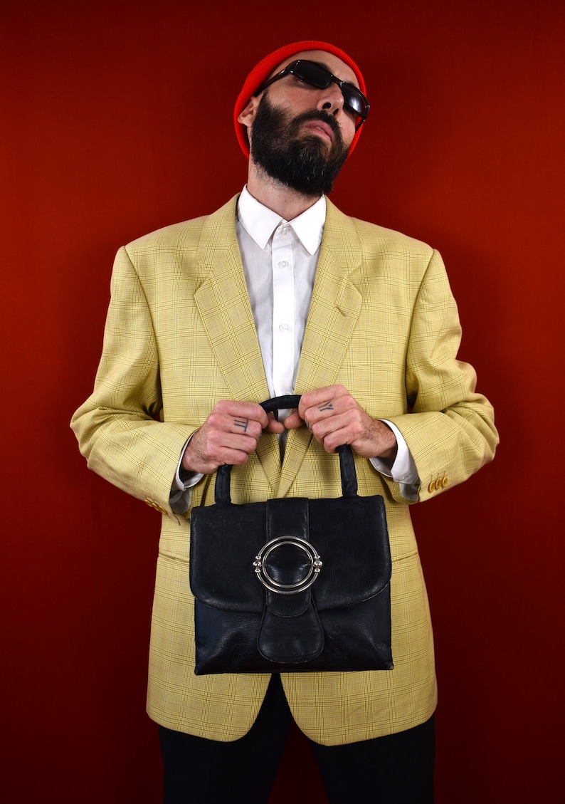 May include: A black leather handbag with a silver-tone circular clasp is held in front of a person wearing a yellow plaid blazer, white shirt, and red beanie. The background is a solid red.