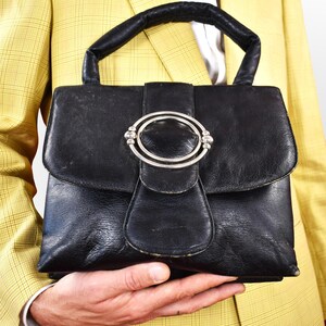 May include: Black leather handbag with a silver-tone clasp. The handbag features a top handle and a flap closure with a decorative oval clasp. The bag is held against a yellow plaid blazer.