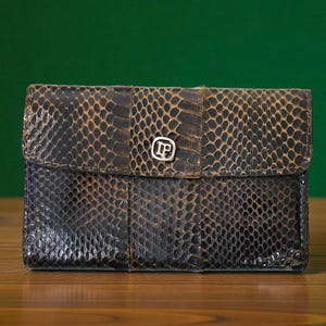 May include: A rectangular, dark brown wallet with a flap closure. The wallet has a snakeskin pattern and a silver-coloured emblem on the flap. The background is a solid green colour.