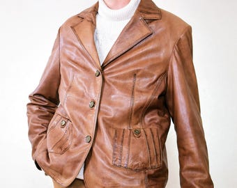 May include: A tan leather jacket with a traditional design. The jacket has a notched collar, button fastening, and several pockets. The model is wearing a white roll-neck sweater and brown corduroy trousers.