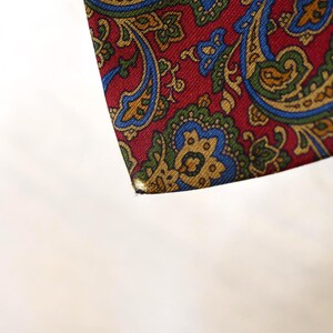 Pu&ograve; includere: Un primo piano di un tessuto con motivo paisley rosso, blu e oro. Il tessuto ha una superficie testurizzata ed &egrave; ripiegato su se stesso.