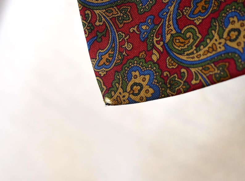 Pu&ograve; includere: Un primo piano di un tessuto con motivo paisley rosso, blu e oro. Il tessuto ha una superficie testurizzata ed &egrave; ripiegato su se stesso.