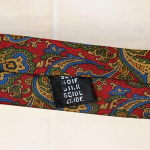 Pu&ograve; includere: Una cravatta di seta con motivo paisley rosso con un'etichetta nera che recita "Bloif Silk Seidel Zurich".