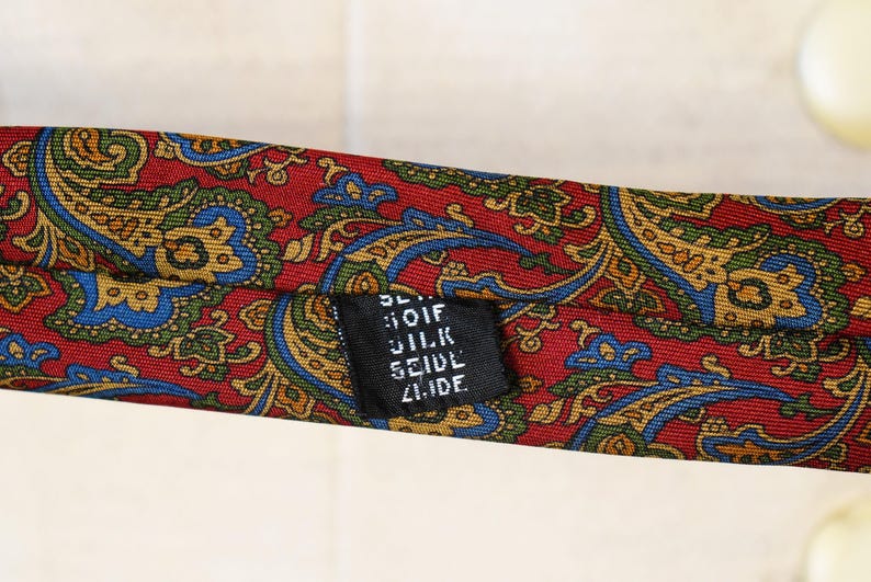 Pu&ograve; includere: Una cravatta di seta con motivo paisley rosso con un'etichetta nera che recita "Bloif Silk Seidel Zurich".