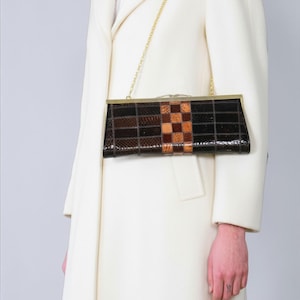 Op de afbeelding: Een rechthoekige clutch met een gouden kettingriem. De clutch heeft een patchwork ontwerp met bruine en bruin/beige vierkanten. Het frame en de sluiting zijn goudkleurig. De clutch wordt tegen een witte jas gehouden.