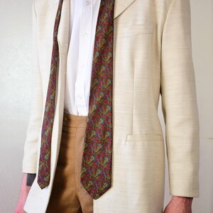 Pu&ograve; includere: Un blazer beige con una cravatta a fantasia paisley. Il blazer ha una chiusura con un solo bottone e una tasca sul lato sinistro. La cravatta &egrave; rossa, blu e verde.