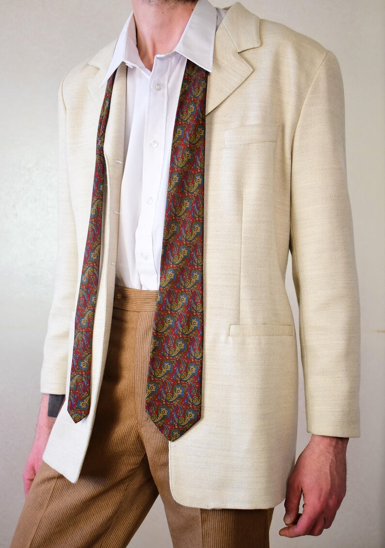 Pu&ograve; includere: Un blazer beige con una cravatta a fantasia paisley. Il blazer ha una chiusura con un solo bottone e una tasca sul lato sinistro. La cravatta &egrave; rossa, blu e verde.