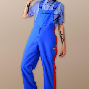 Könnte beinhalten: Blaue Latzhose mit roten Seitenstreifen und Reißverschluss. Die Latzhose hat eine einzelne Tasche auf der Vorderseite. Die Person trägt ein hellblaues Hemd mit Knöpfen und eine rote Mütze.