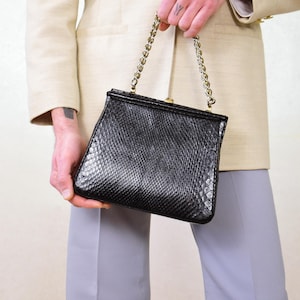 Peut inclure: Un sac à main noir texturé avec une chaîne argentée. Le sac est de forme rectangulaire avec une fermeture à fermoir. La personne porte un blazer beige et un pantalon gris.