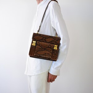 Könnte beinhalten: Eine rechteckige Umhängetasche mit braun-schwarzem Schlangenledermuster. Die Tasche verfügt über eine Fronttasche mit Klappe, ein Hauptfach mit Überschlag und goldfarbene Metallverschlüsse. Sie wird mit einem dünnen braunen Schulterriemen getragen.