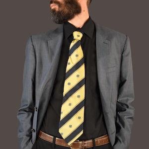 Puede incluir: Un hombre con una chaqueta de traje gris, una camisa negra y una corbata a rayas amarilla y azul. Lleva gafas de sol y tiene barba.