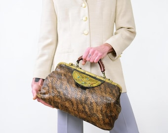 May include: A brown and black textured leather handbag with a brass frame and a decorative brass plate featuring small blue and red accents. The bag has a dark brown handle and a metal clasp closure, showcasing a distinct mottled pattern throughout.