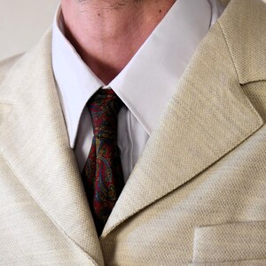 Pu&ograve; includere: Un blazer beige in lino con una camicia bianca con colletto e una cravatta con motivo paisley. La cravatta &egrave; rossa, blu e gialla.